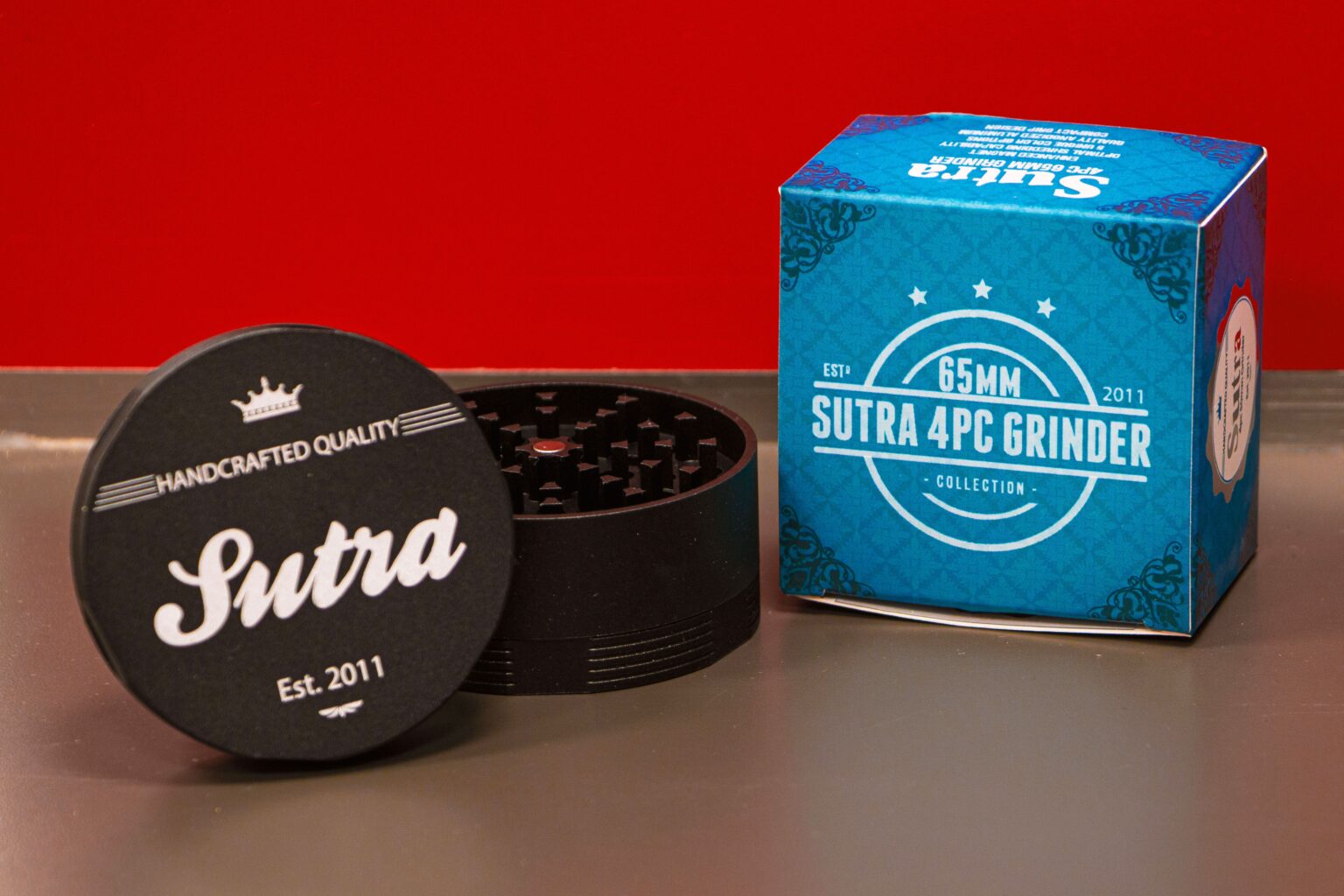 Black 65mm Sutra four piece grinder open beside its blue retail box on a grey surface